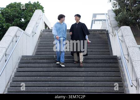 Verschiedene männliche Freunde, die eine breite Steintreppe auf dem Campus hinunterlaufen, Abitur im Kleid mit gerolltem Diplom Stockfoto