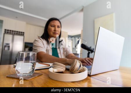 Asiatische Frau, die am Küchentisch mit grünem Oberteil, Kopfhörern und Mikrofon in der Nähe tippt Stockfoto