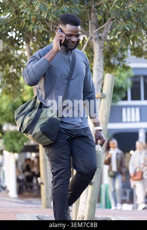 Erwachsener afrikanischer Mann mit grauem Oberteil, der auf einem von Bäumen gesäumten Bürgersteig läuft und am Telefon mit Seetasche spricht Stockfoto