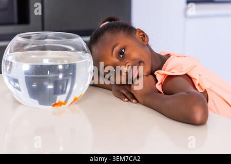 Weibliches Kind lehnt sich auf eine glänzende Arbeitsplatte in der Küche und beobachtet Fischschalen mit zwei orangefarbenen Goldfischen Stockfoto