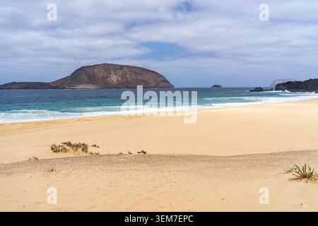 Eine ruhige Strandszene auf La Graciosa, die die wunderschöne Sandküste, das türkisfarbene Wasser und die entfernte Silhouette der Insel zeigt. Stockfoto