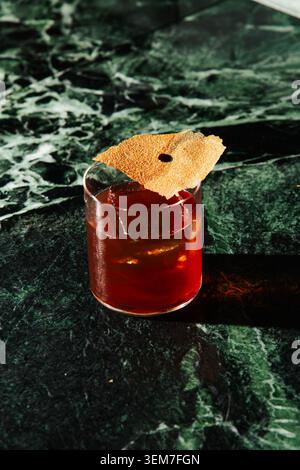 Raffinierter roter Cocktail mit klarem Eiswürfel und knuspriger Verzierung auf einem dunkelgrünen Marmortisch. Elegantes Mixologiekonzept mit hartem Lichtschatten. Stockfoto