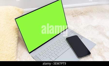 Laptop-Modell mit leerem grünem Bildschirm neben einem Smartphone auf einem gemütlichen Pelzteppich, perfekt für kreatives Design und digitale Präsentationen Stockfoto