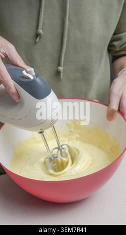 Die Person hält einen elektrischen Handmixer, während sie gelben Teig in einer roten Rührschüssel auf einer Küchenarbeitsfläche mit einem neutralen Weichfokus mischt Stockfoto