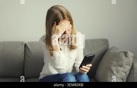 Traurige aufgebrachte, schreiende junge Frau, die das Telefon hält und unter Stress leidet, nachdem sie emotionale Nachrichten gelesen hat, die auf der Couch zu Hause sitzen Stockfoto