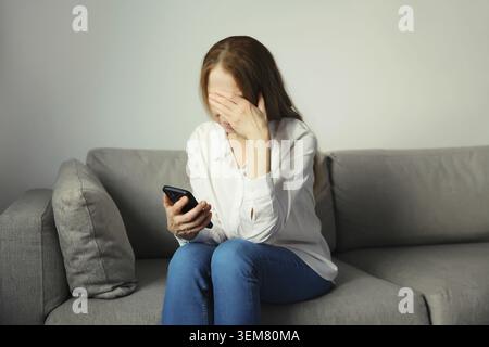 Traurige aufgebrachte, weinende Frau, die das Telefon hält und unter Stress leidet, nachdem sie emotionale Nachrichten gelesen hat, die auf der Couch zu Hause sitzen Stockfoto