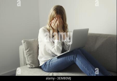 Traurige Verärgerung überarbeitete junge Frau, die unter Kopfschmerzen, Stress und Migräne leidet, während sie mit einem Laptop arbeitet, der auf der Couch im Wohnzimmer sitzt Stockfoto