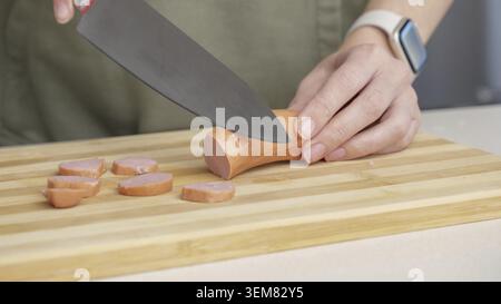 Nahaufnahme einer Frau, die Wurst auf einem hölzernen Schneidebrett in einer Küche mit verschiedenen Zutaten schneidet Stockfoto