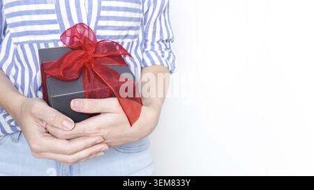 Eine junge Frau lächelt, während sie eine wunderschön verpackte Geschenkbox mit einem roten Band hält. Sie trägt ein gestreiftes Hemd und Jeans und sieht glücklich aus. Der bac Stockfoto