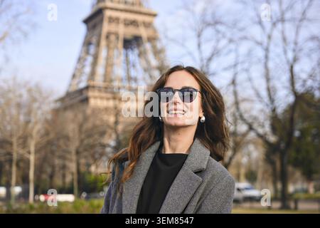 Attraktive junge Frau mit langen Haaren, Sonnenbrille und grauem Mantel, die an einem sonnigen Frühlingstag vor dem Eiffelturm lächelt Stockfoto