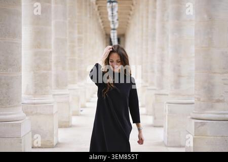 Fröhliche junge Frau in schwarzem Kleid, die selbstbewusst durch die klassische architektonische Kolonnade im Palais Royal in Paris, Frankreich, wandert Stockfoto