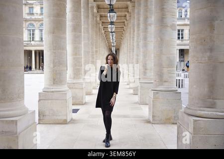 Selbstbewusste Frau in schwarzem Kleid, die durch die elegante Steinkolonnade des Palais Royal in Paris, Frankreich, geht. Stilvolle urbane Lifestyle-Szene Stockfoto