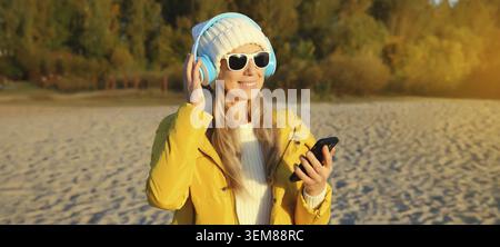 Glückliche inspirierte Frau mit Telefon, die Musik im Kopfhörer draußen an der Herbstküste am Meer hört, Bäume mit gelben Blättern, Mädchen-Blick Stockfoto