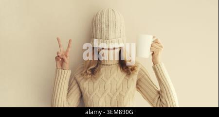 Glückliche lustige junge Frau mit einer Tasse Kaffee trinken im Winter gestrickte Hut, Pullover, der Spaß auf beigem Hintergrund hat Stockfoto