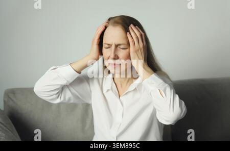 Erschöpfte müde Frau, die den Kopf berührt, unter Kopfschmerzen, Stress, Müdigkeit, Schläfenschmerzen, Migräne, lautes Geräusch, Spannung auf der Couch zu Hause Stockfoto