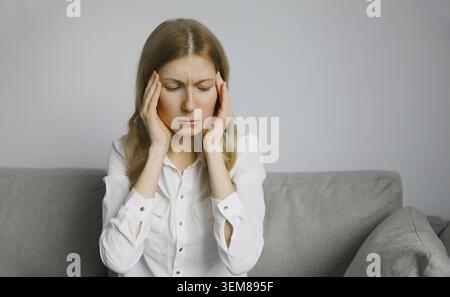 Erschöpfte, müde junge Frau, die den Kopf berührt, unter Kopfschmerzen, Stress, Müdigkeit, Schläfenschmerzen, Migräne, lautes Geräusch, Spannung auf der Couch Stockfoto
