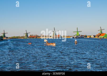 Amsterdam, Niederlande, 22.04.2026 - traditionelle niederländische Windmühlen im Dorf Zaanse Schans neben dem Fluss mit Touristen in einem Ruderboot unter freiem Himmel Stockfoto