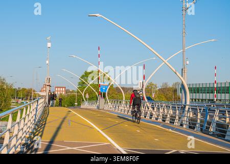 Amsterdam, Niederlande, 22.04.2026 - Radfahrer fahren auf einem Radweg über moderne Brücke mit futuristischen Straßenlaternen an sonnigen Tagen Stockfoto