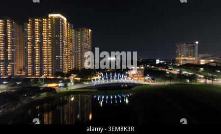 Blick aus der Vogelperspektive auf Summarecon Bekasi bei Nacht mit beleuchteten Hochhausapartments und einer leuchtenden Brücke, die auf dem Wasser in Kecamatan Medan S reflektiert Stockfoto