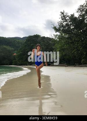 Schöne junge Frau in einem blauen Badeanzug, die fröhlich auf die Kamera entlang eines tropischen Strandes läuft, lächelt und den Moment genießt. Dynamischer Lifestyle Stockfoto
