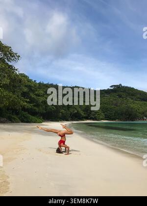 Flexible junge Frau, die eine elegante Kopfstand-Yoga-Pose an einem unberührten tropischen Strand spielt, wobei ein Bein gebeugt und das andere voll ausgestreckt ist Stockfoto