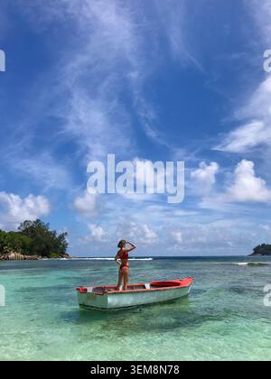 Schlanke Frau von hinten in einem roten Bikini, anmutig auf einem traditionellen Fischerboot, das im flachen kristallklaren türkisfarbenen Wasser schwimmt. Stockfoto