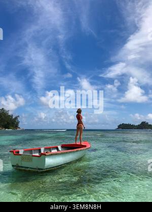 Schlanke Frau von hinten in einem roten Bikini, anmutig auf einem traditionellen Fischerboot, das im flachen kristallklaren türkisfarbenen Wasser schwimmt. Stockfoto