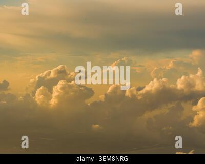 Flauschige goldene Cumuluswolken, die sich am Abendhimmel erheben, umrahmen ein winziges kommerzielles Flugzeug, das hoch über einem dunklen, fernen Horizont fliegt. Stockfoto