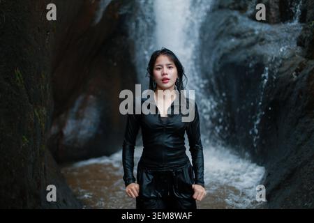 Selbstbewusste junge Frau, die vor einem wunderschönen Wasserfall steht, umgeben von dunklem Felsen in der Natur, mit schwarzer nasser Kleidung, die intensiv aussieht Stockfoto