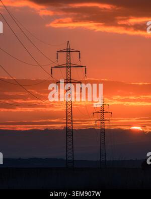 Blick auf Hochspannungsleitungen und Strommasten vor einem leuchtenden orangefarbenen Sonnenuntergangshimmel mit verstreuten Wolken Kyjov, Südmährische Re Stockfoto