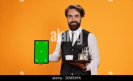 Der freundliche Kellner zeigt ein Tablet mit Greenscreen auf der Kamera und zeigt eine leere Vorlage für ein Copyspace-Modell. Erfahrener Butler in Anzug Haltevorrichtung mit isoliertem leerem Display und Essenstablett. Stockfoto