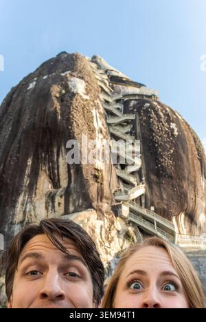 Ein Mann und eine Frau stehen zusammen im Vordergrund und lächeln in die Kamera, mit einer hohen Felsklippe und einer Metalltreppe, die in den Felsen gehauen ist Stockfoto
