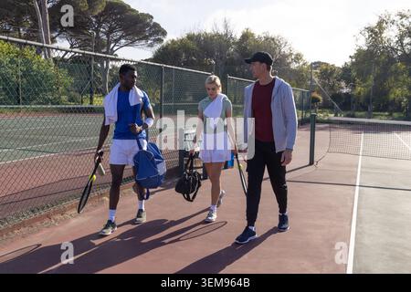 Verschiedene Freunde, die auf einem Lehmpassweg laufen, mit Tennisschlägern, Taschen, Handtüchern und sichtbarer Bank Stockfoto
