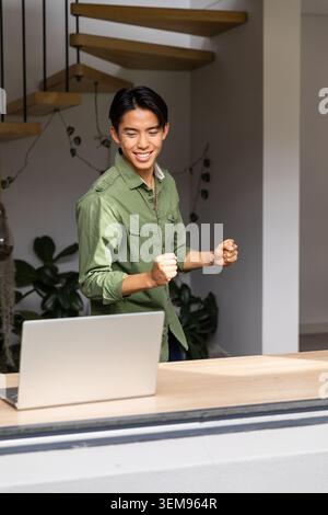 Asiatischer Mann in grünem Hemd, dunkle Hosen, die hinter Holztheke stehen und die Fäuste in Richtung geöffnetem Laptop heben Stockfoto
