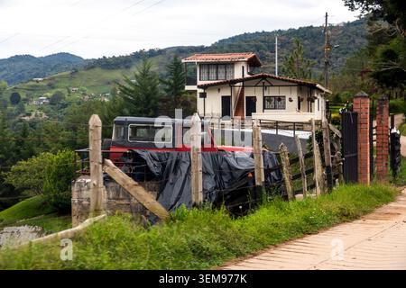 Ein kleines Haus auf einem grünen Hügel mit einem Pickup-Truck, der hinter einem rustikalen Zaun geparkt ist, umgeben von sanften Bergen, Bäumen und einem gepflasterten Bürgersteig Stockfoto