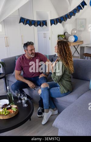 Ein Paar sitzt auf einem grauen Sofa im Wohnzimmer, spricht über Wein, Käsebrett unter Geburtstagsbanner Stockfoto