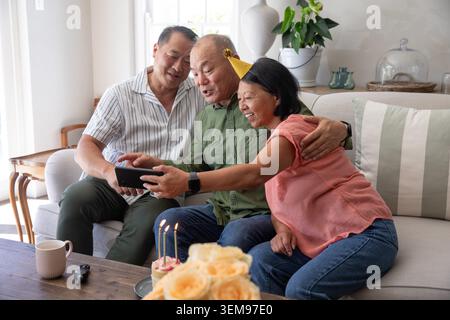 Asiatische Familie, ältere Mutter und Söhne, die sich zu Hause auf das Sofa lehnen, Smartphone und Kuchen teilen Stockfoto