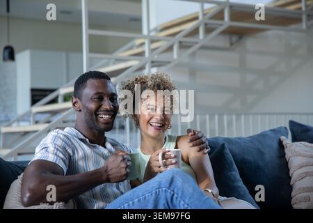 Afroamerikanisches Paar, das auf einem dunkelblauen Sofa im modernen Wohnzimmer sitzt und Keramikbecher hält Stockfoto