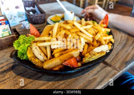 Ein großer schwarzer Teller mit goldenen Pommes frites, gegrilltem Fleisch, Salat und Tomaten auf einem Holztisch Stockfoto