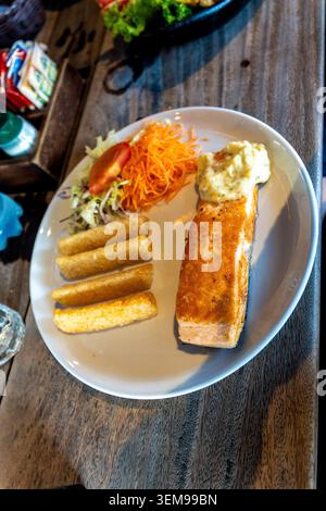 Ein überzogenes Lachsfilet mit cremigem Belag, drei goldenen frittierten Beilagen und ein kleiner frischer Salat, serviert auf einem weißen Teller über einem rustikalen Holztisch Stockfoto