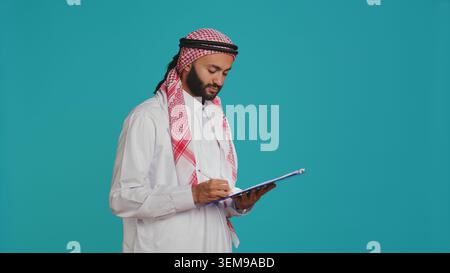 Arabische Person, die einen Brief auf Akten schreibt, über blauem Hintergrund vor der Kamera steht, an einem neuen Zeitplan arbeitet und Notizen macht. Ein Typ in islamischer Kleidung, der sich die Checkliste für Papiere im Studio ansieht. Stockfoto