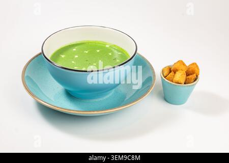 Eine lebendige grüne Gemüsepüree-Suppe mit cremefarbenen Herzen, serviert in einer stilvollen türkisfarbenen Keramikschale neben knusprigen goldenen Croutons Stockfoto