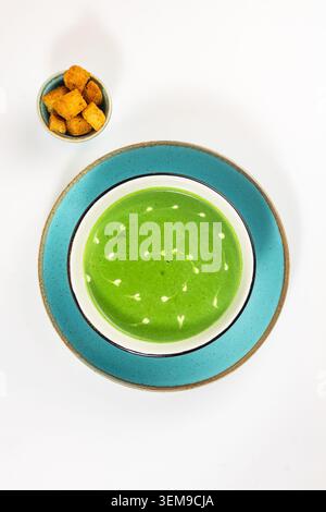Minimalistisch geometrischer Blick von oben auf eine üppige grüne Spinat-Creme-Suppe, kombiniert mit einer kleinen Schüssel knuspriger Croutons vor einem nahtlosen weißen Hintergrund Stockfoto