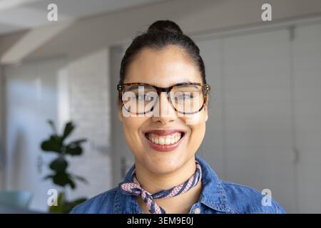 Frau mit Schildkrötenbrille, Denim-Hemd, Halstuch, Lächeln zu Hause in der Nähe der Pflanze, weiße Türen Stockfoto