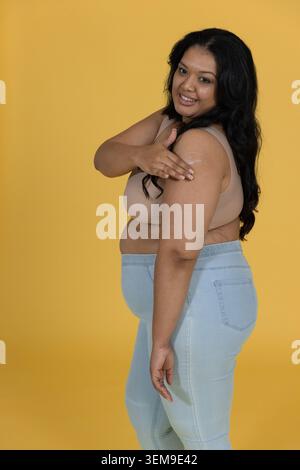 MittelErwachsene Frau in gelbem Studio mit beigem BH und Jeans und weißer Creme auf den rechten Arm Stockfoto