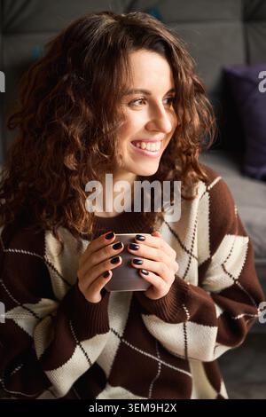 Entspannte junge Frau, die heißes Getränk drinnen genießt, steht für Komfort, langsames Leben, emotionales Wohlbefinden und modernen Lebensstil Stockfoto