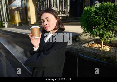 Porträt einer brünetten Frau in eleganter schwarzer Oberbekleidung, die draußen an der Fassade des städtischen Gebäudes Kaffee genießt. Ideal für Lifestyle-Werbung, Street-Fashion-Werbung, saisonale Inhalte und Branding Stockfoto