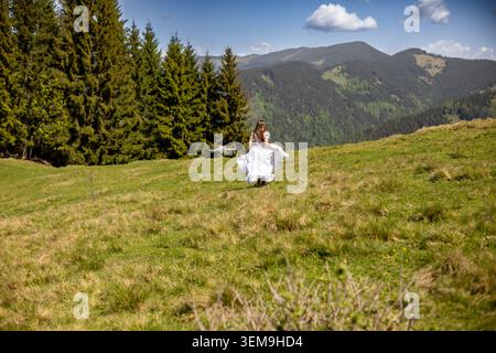 Frau, die mit Drohne auf einer Bergwiese läuft. Eine junge Frau in fließendem weißem Kleid rennt über eine sonnige Bergwiese, während eine Drohne über die Landschaft fliegt Stockfoto