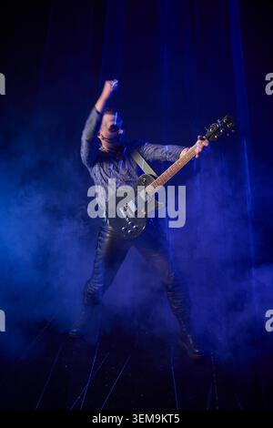 Erwachsener männlicher Musiker mit dunklem Haar, der leidenschaftlich schwarze E-Gitarre auf einer rauchgefüllten Bühne unter blauer Beleuchtung spielt, dynamisch erhobener Faust und Leath Stockfoto