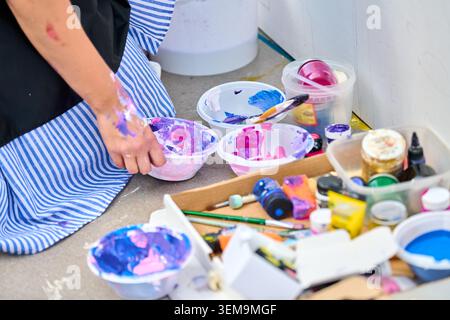 Nahaufnahme eines Künstlers, der leuchtend rosa und blaue Acrylfarbe in Schüsseln auf dem Boden mit verstreuten Farbtuben, Pinseln und Gläsern gegen weiße Wand rührt Stockfoto